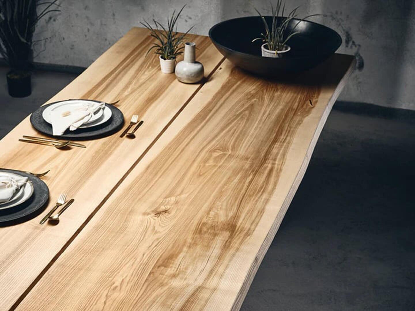 Ein moderner Esstisch aus Holz mit einer natürlichen Kante, gedeckt mit zwei Gedecken, goldenem Besteck, einem schwarzen Teller und einer weißen Serviette. Dekorative Pflanzen und eine schwarze Schale sind als Mittelstück arrangiert.