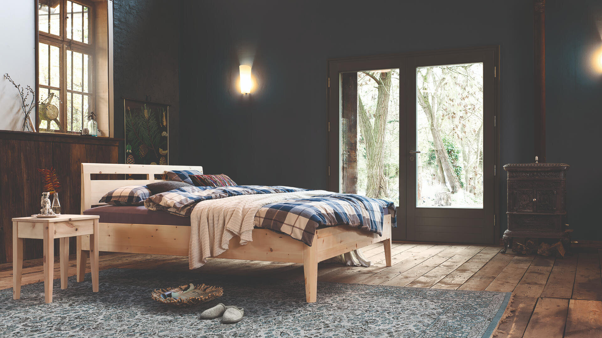 Gemütliches Schlafzimmer mit einem Holzbett, karierter Bettwäsche und einem kleinen Nachttisch mit einer Vase. Das Zimmer hat Holzböden, einen verzierten Teppich, einen Holzofen und große Glastüren, die den Blick auf die Bäume draußen freigeben.