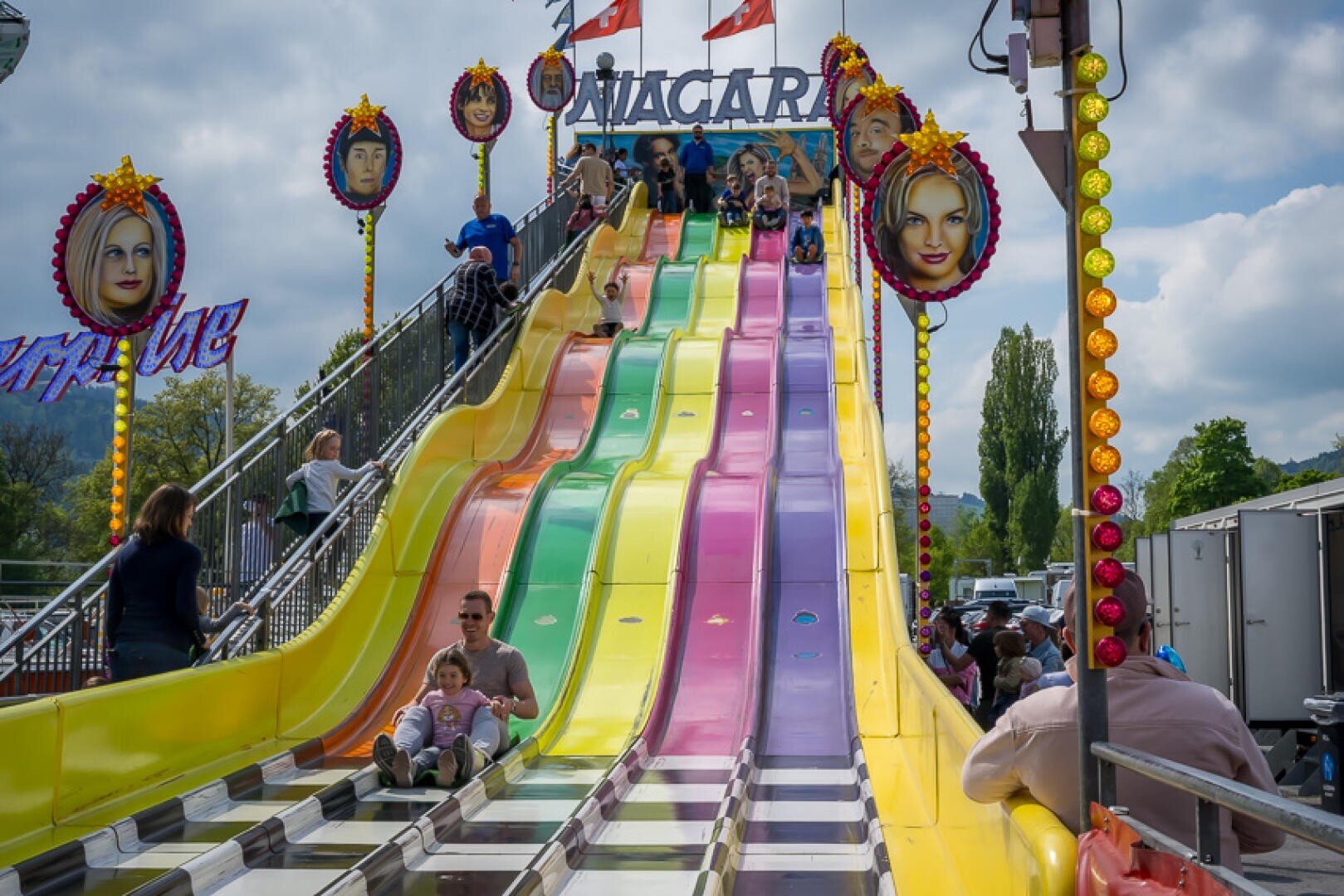 Menschen und Kinder rutschen auf einer farbenfrohen, wellenförmigen Rutsche mit dem Namen Niagara hinunter, die auf jeder Seite mit festlichen Lichtern und großen Gesichtern geschmückt ist. Der Himmel ist teilweise bewölkt und im Hintergrund sind Bäume zu sehen.