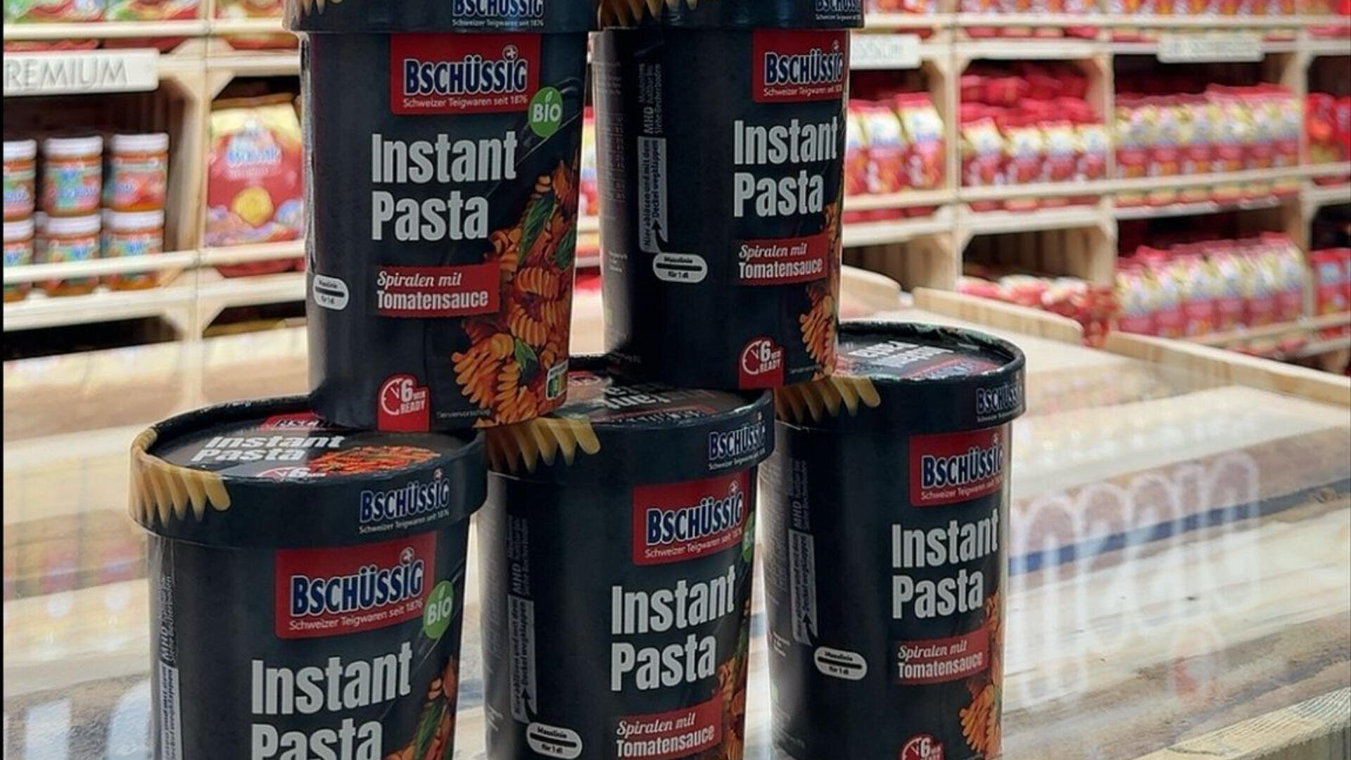 Fünf schwarze Becher Bschüssig Instant Pasta in Tomatensoße stehen pyramidenförmig auf einer Ladentheke, im Hintergrund sind Regale mit verpackten Lebensmitteln zu sehen.