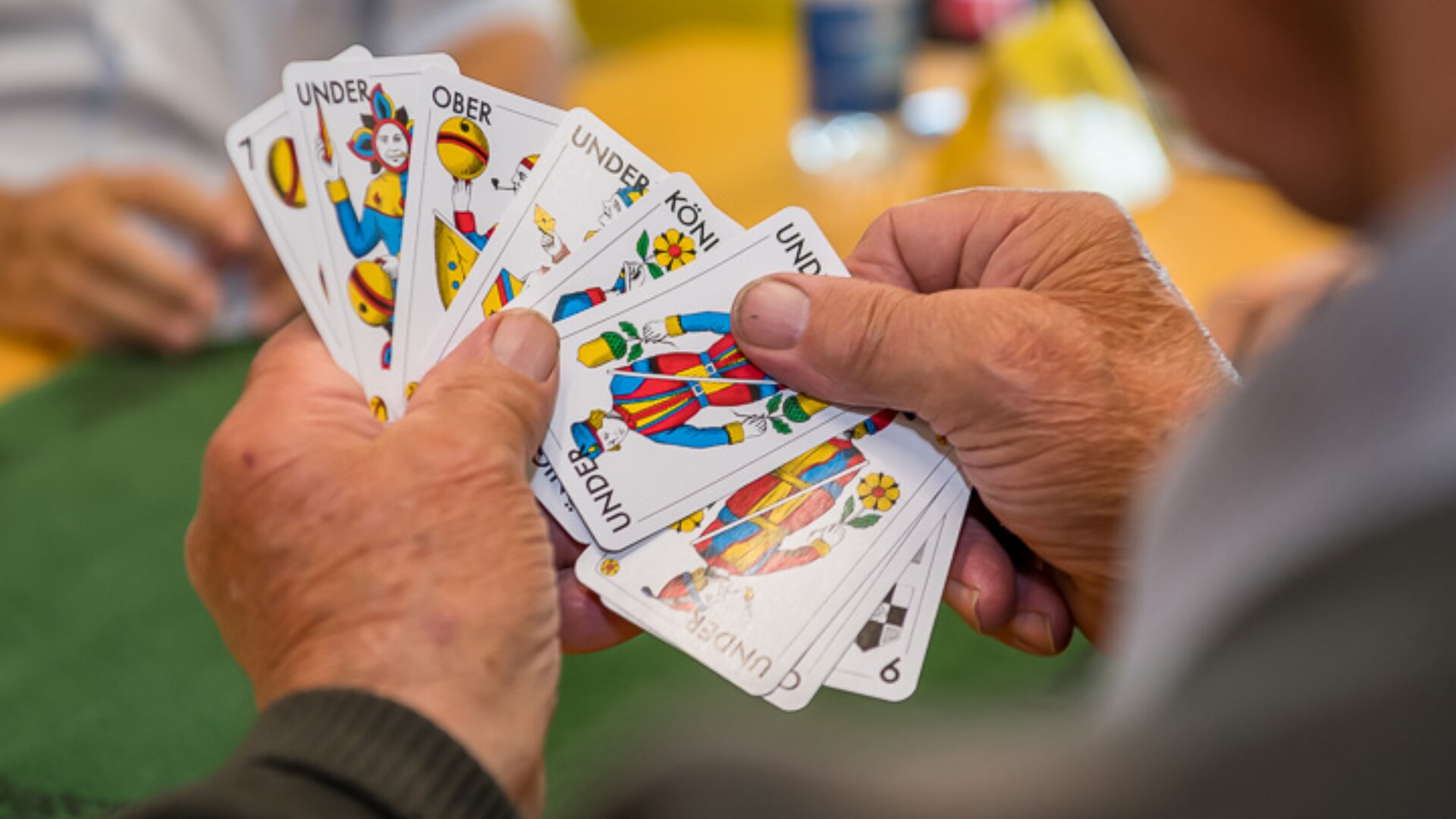 Eine Person hält einen Fächer mit bunten Spielkarten in den Händen, auf denen verschiedene Zahlen, Nummern und Symbole zu sehen sind. Die Szene erinnert an ein Kartenspiel, das an einem Tisch gespielt wird.