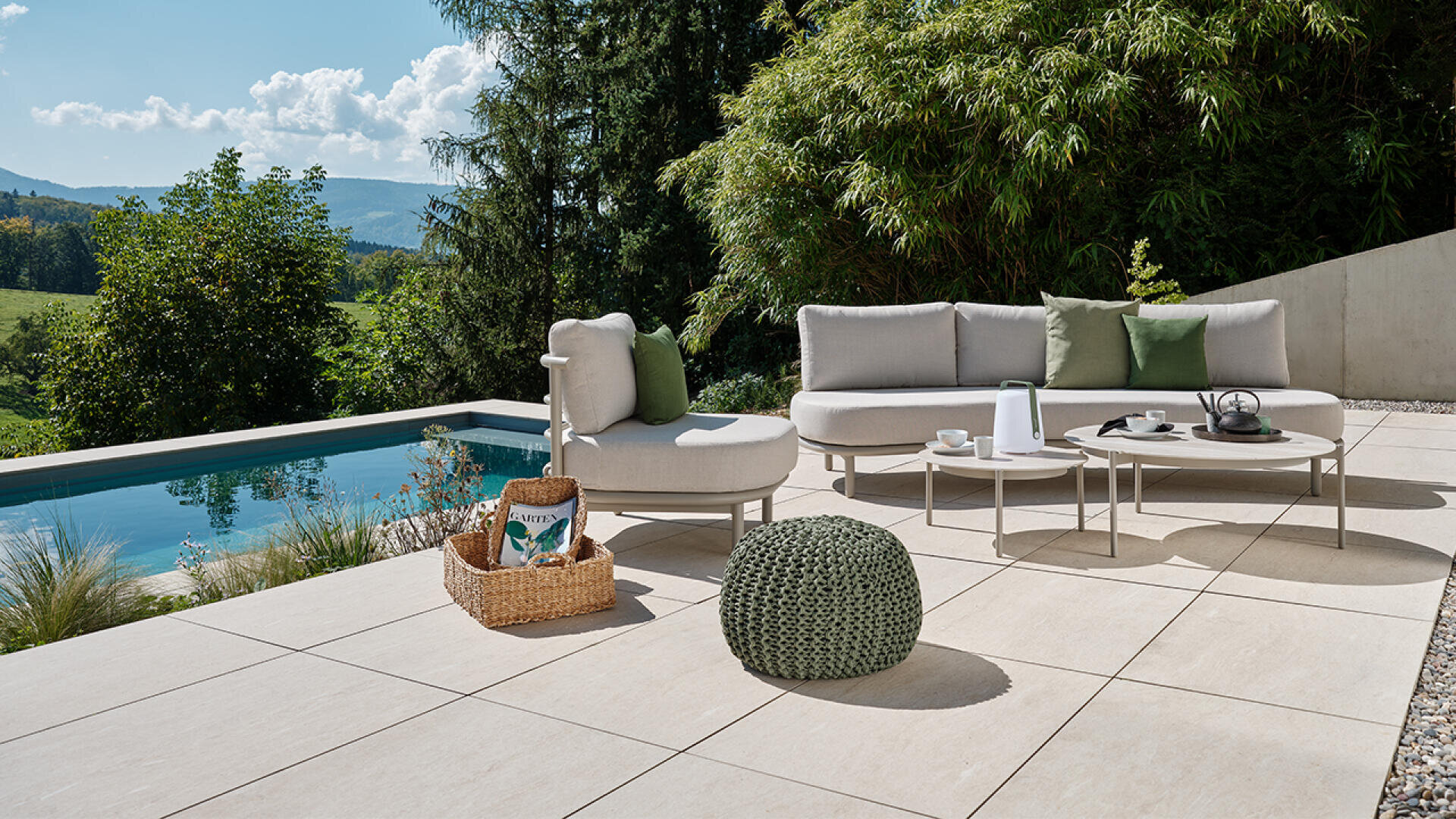 Moderne Außenterrasse mit hellen Fliesen, einem geschwungenen grauen Sofa, einem runden grünen Hocker, Couchtischen mit Teekanne und Tassen, Korb und Pool; umgeben von üppigem Grün und fernen Bergen.