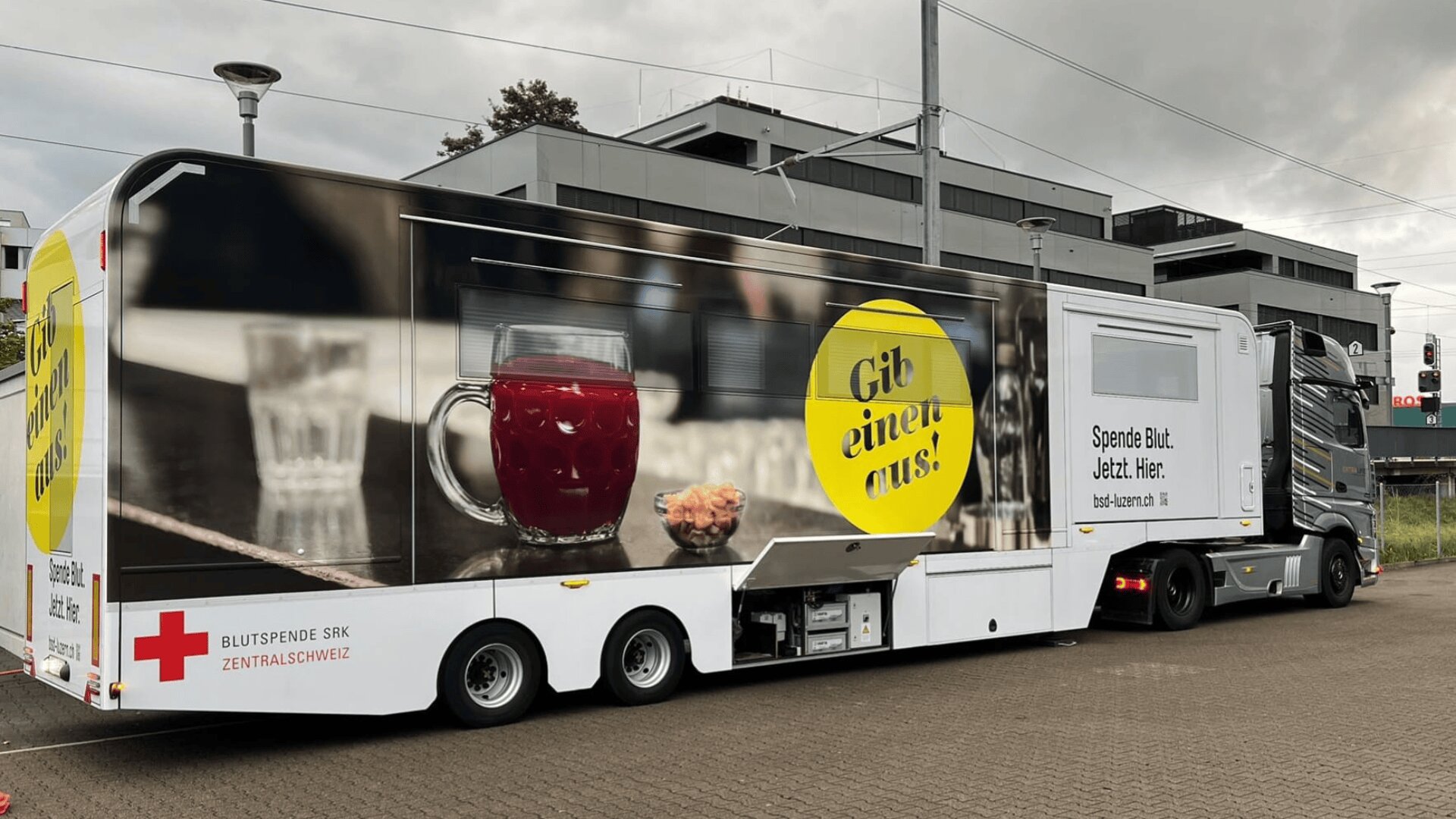 Ein großer Blutspendetransporter, der vor einem modernen Gebäude geparkt ist, mit einer Grafik eines Glases mit roter Flüssigkeit, Gebäck und fettgedrucktem deutschen Text, der für Blutspenden wirbt. Das Logo des Schweizerischen Roten Kreuzes ist auf dem Anhänger zu sehen.