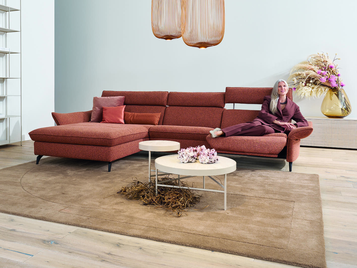 Eine Frau in einem lilafarbenen Anzug lehnt sich auf einem modernen roten Sektionssofa in einem stilvollen Wohnzimmer mit hellem Holzboden, einem beigen Teppich, runden Couchtischen und dekorativen Vasen mit Blumen zurück.