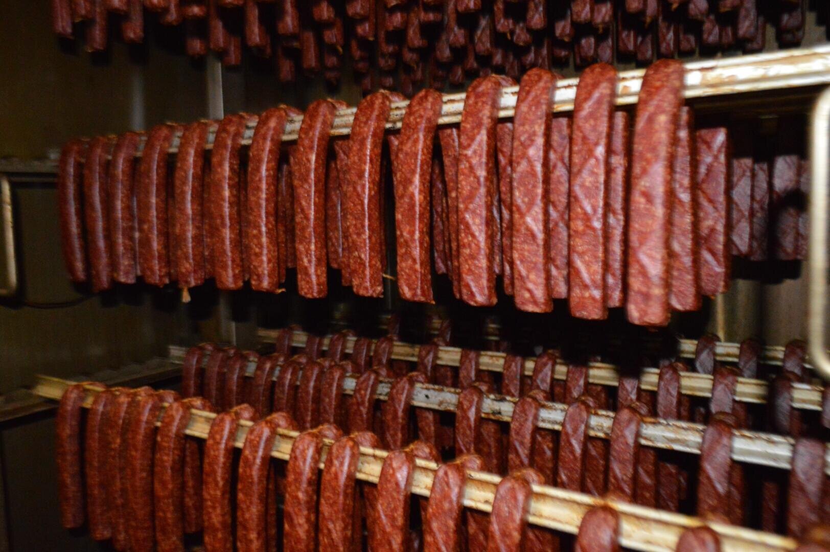 Reihen von Wurststangen, die senkrecht auf Metallgestellen hängen, wahrscheinlich in einer Räucherkammer oder einem Trockenraum, mit einem unscharfen Hintergrund, der weitere Würste zeigt.