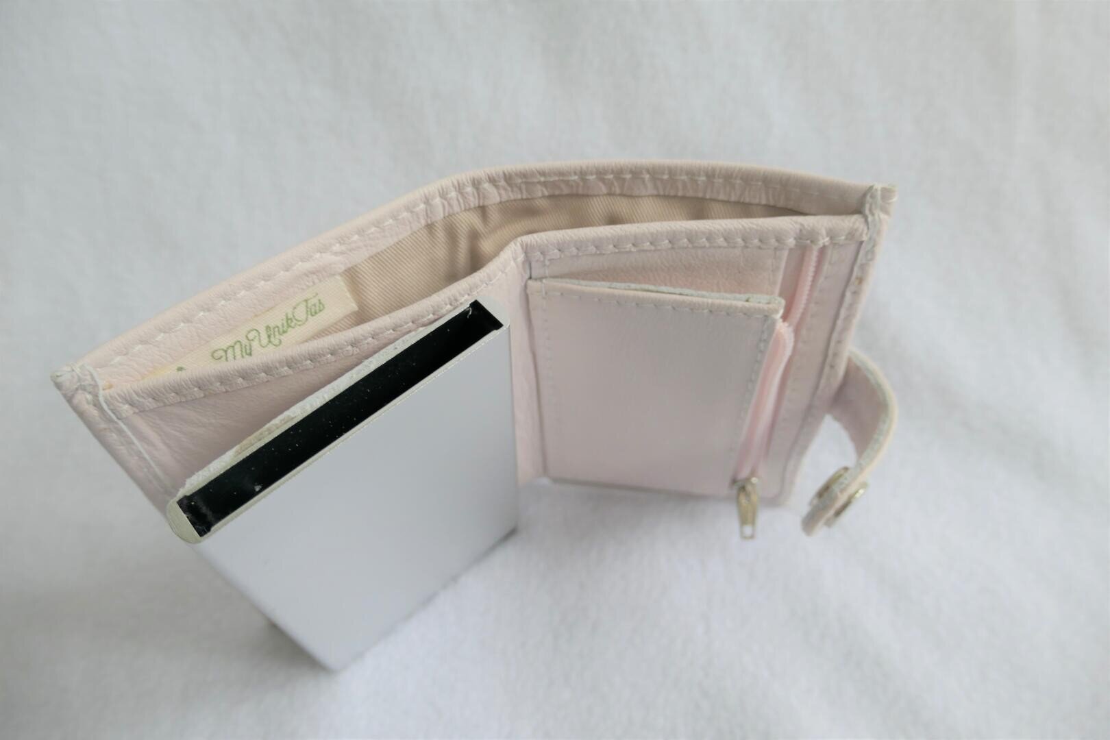Eine hellrosa Brieftasche, teilweise geöffnet, mit einem Münzfach mit Reißverschluss und einem kleinen silbernen rechteckigen Spiegel im Inneren. Das Portemonnaie ruht auf einer weißen Fläche.
