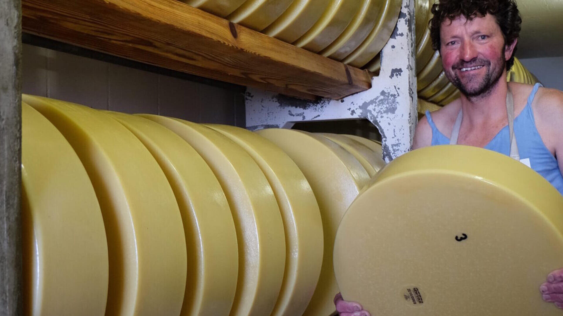 Ein lächelnder Mann hält einen großen Laib Käse in einem Lagerraum, der mit gestapelten Käselaiben auf Holzregalen gefüllt ist.