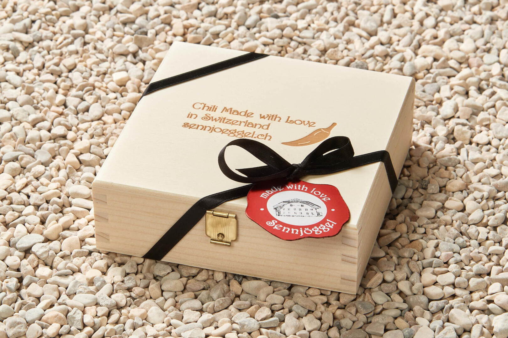 Eine hölzerne Geschenkbox mit schwarzem Band und Verschluss sitzt auf kleinen Kieselsteinen. Auf dem Deckel ist "Chili Made with Love in Switzerland sennjogsglch." eingraviert. Auf einem roten Anhänger steht "made with love sennjogsglch.".