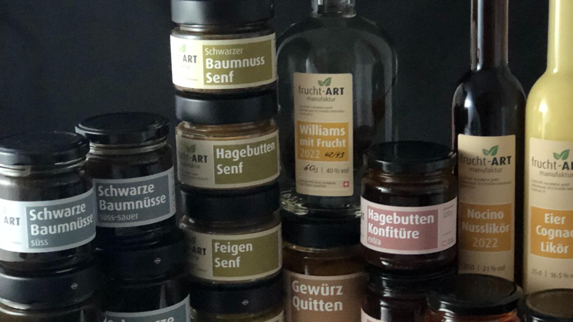 Eine Vielzahl von etikettierten Gläsern und Flaschen mit Brotaufstrichen, Senf, Likören und Konfitüren sind nebeneinander angeordnet und tragen deutsche Produktnamen wie Schwarze Baumnuss Senf, Hagebutten Senf und Williams mit Frucht.