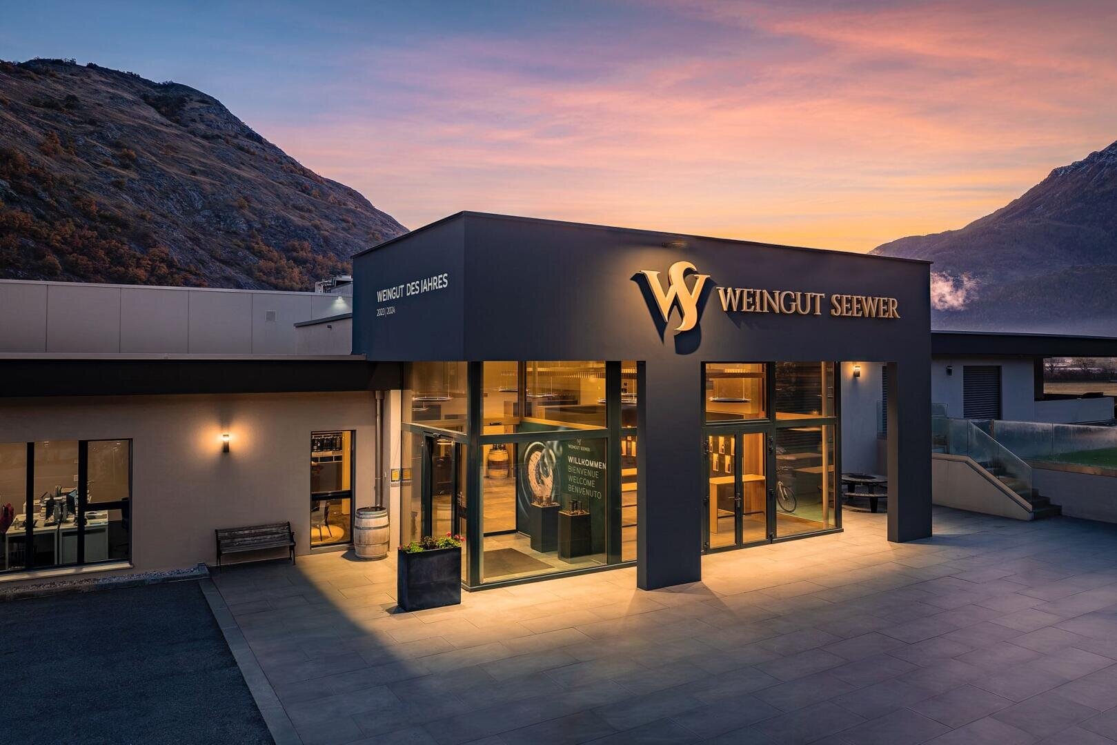 Modernes Kellereigebäude mit der Bezeichnung Weingut Seewer bei Sonnenuntergang, mit großen Glasfenstern und warmer Innenbeleuchtung, umgeben von Bergen und einem klaren Himmel mit bunten Wolken.