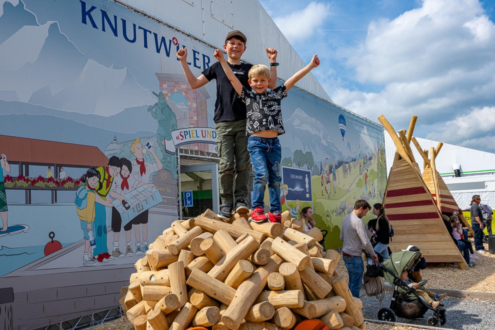 Zwei Kinder stehen mit aufgeregt erhobenen Armen auf einem Stapel großer Holzstämme. Um sie herum stehen Menschen und Familien, und im Hintergrund sind ein Wandgemälde und ein Spielplatz zu sehen.