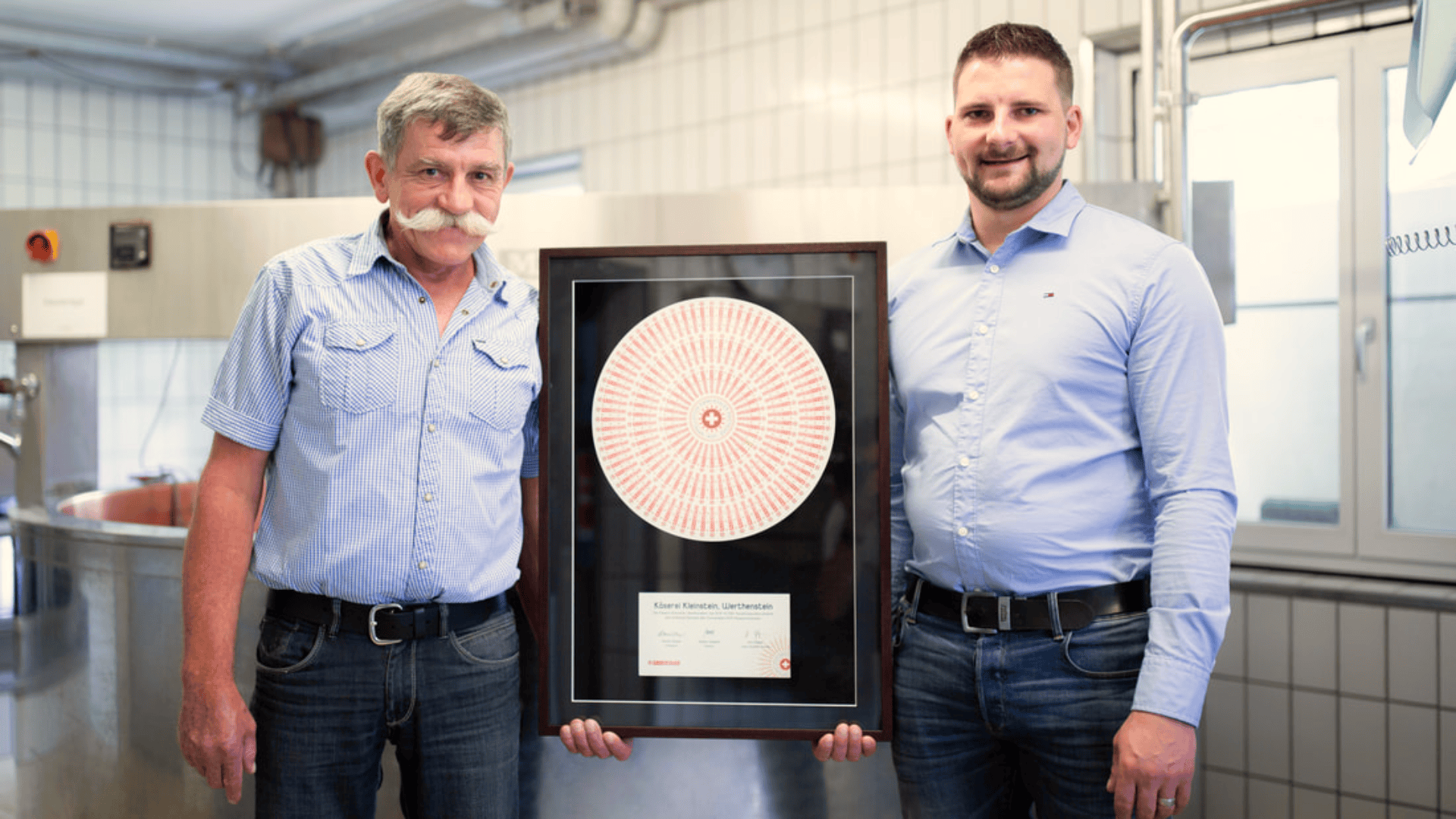 Zwei Männer stehen in einem Haus und halten eine gerahmte Urkunde mit einem kreisförmigen rot-weißen Muster. Beide lächeln und tragen blaue Hemden und Jeans. Der Hintergrund zeigt eine geflieste Wand und Industrieanlagen.