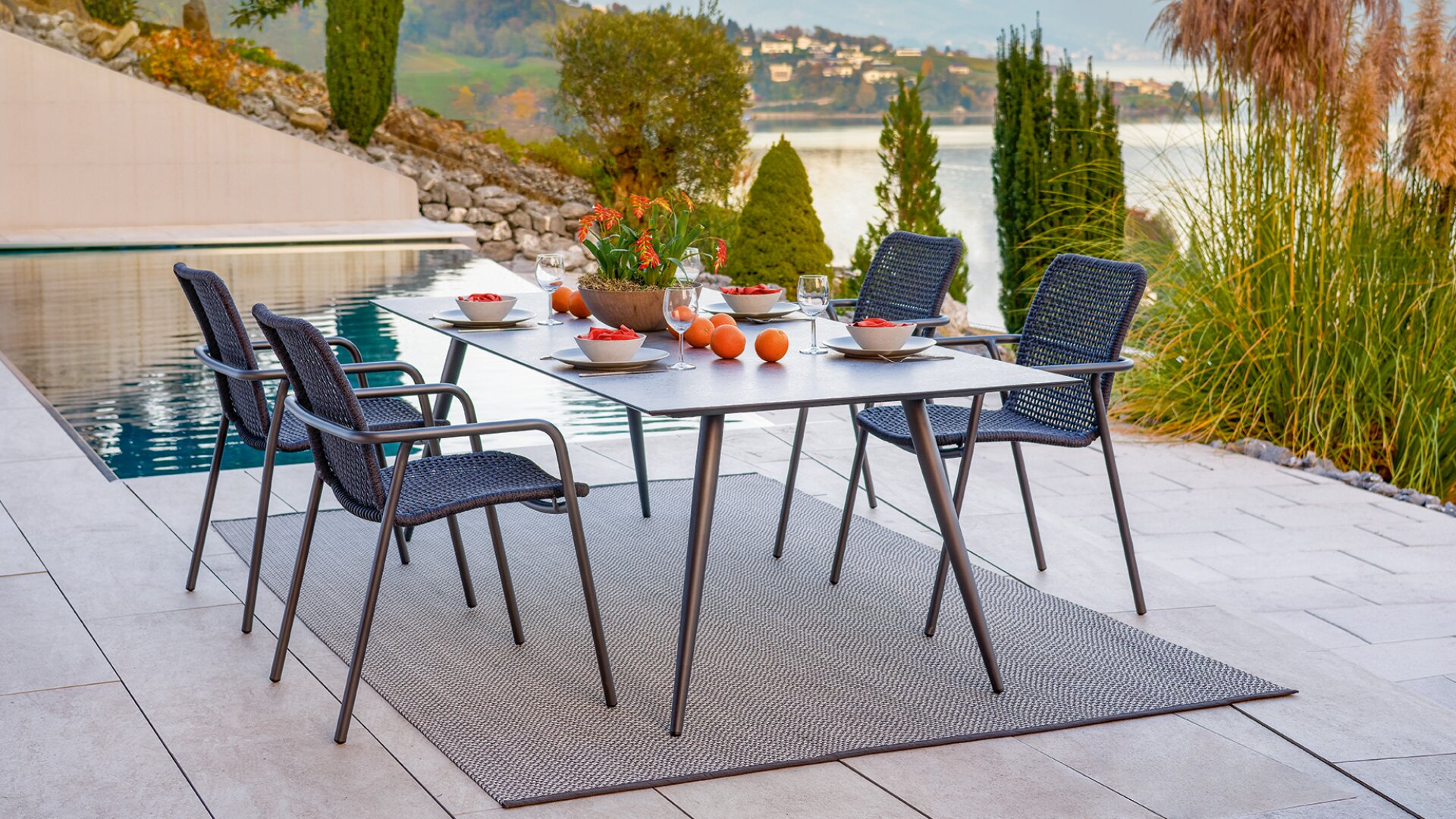 Ein moderner Esstisch für vier Personen im Freien mit Obstschalen, Orangen und Wassergläsern auf einer Terrasse neben einem Pool, mit Blick auf einen malerischen See und die Hügel.