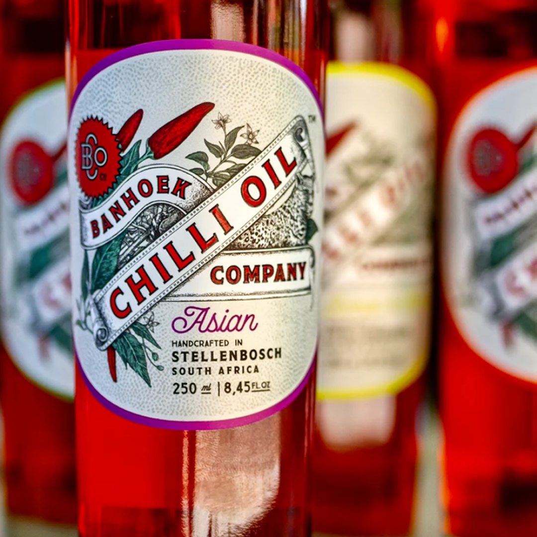 Nahaufnahme einer Flasche mit der Aufschrift Banhhoek Chilli Oil Company, Asian, mit Chilischoten und Blättern auf dem Etikett. Die Flasche enthält rotes Chili-Öl und wird in Stellenbosch, Südafrika, in Handarbeit hergestellt.