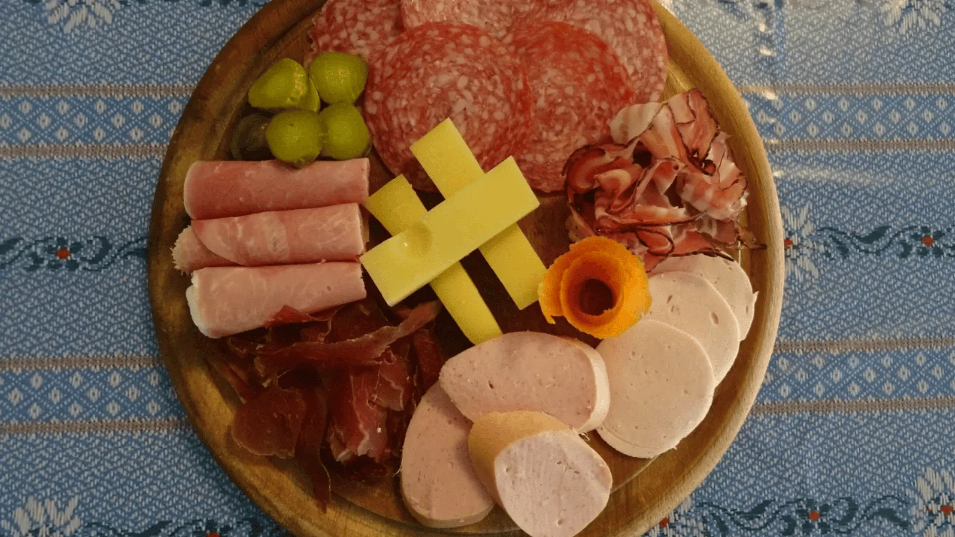 Eine Holzplatte mit verschiedenen Aufschnittarten, darunter Salami, Schinken, Bresaola, Käsescheiben, grüne Weintrauben und eine Scheibe Karotte auf einer blau gemusterten Tischdecke.