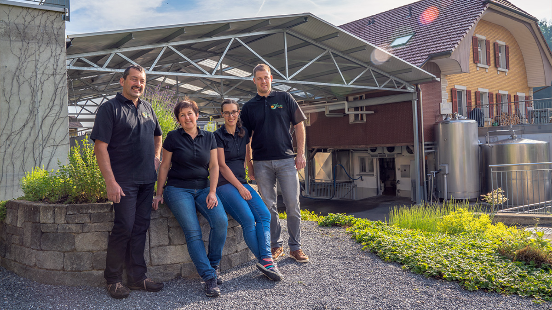 Vier Personen in schwarzen Hemden stehen und sitzen an einem sonnigen Tag vor einem Gebäude mit Metalltanks und einer Gewächshauskonstruktion zusammen und lächeln in die Kamera. Im Vordergrund sind Grünflächen und ein Garten zu sehen.