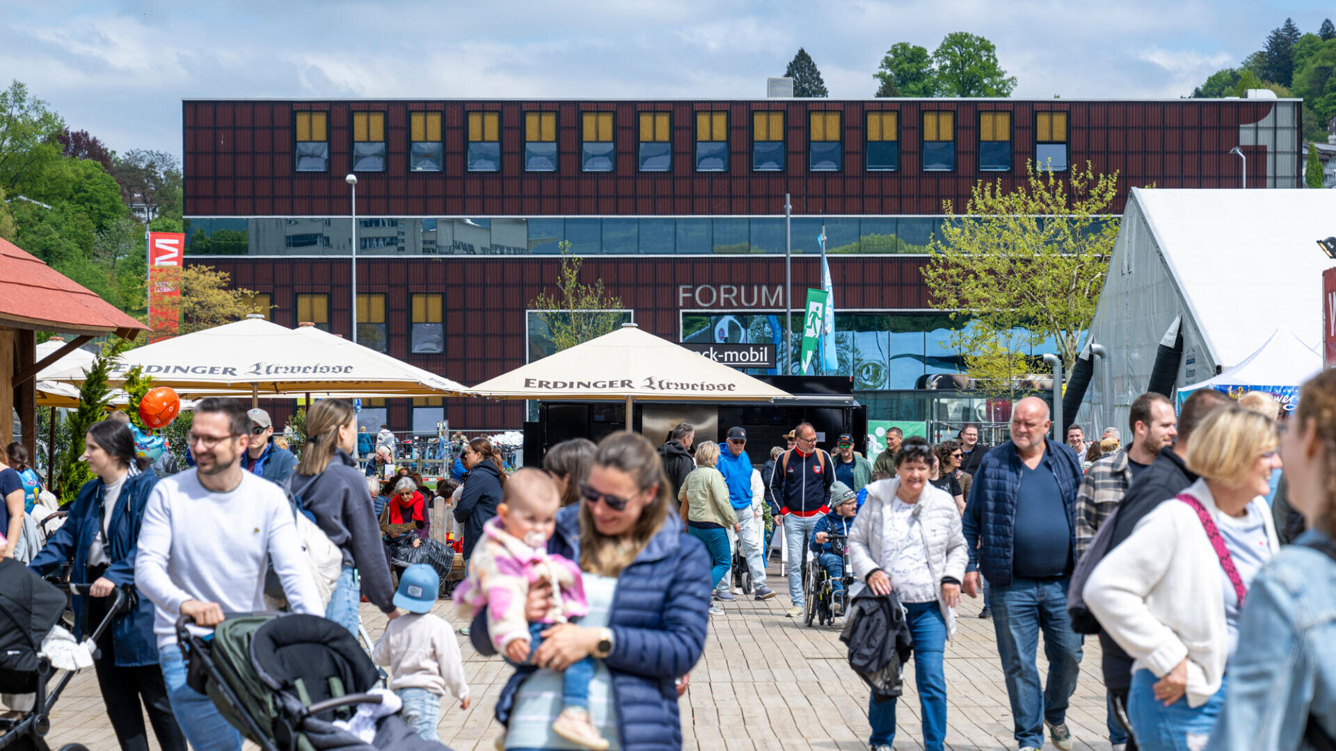 Eine Menschenmenge, darunter Familien mit Kindern und Kinderwagen, spaziert an einem sonnigen Tag im Freien vor einem modernen Gebäude mit dem FORUM-Schriftzug an der Fassade und großen weißen Sonnenschirmen. Im Hintergrund sind Bäume und Grünflächen zu sehen.
