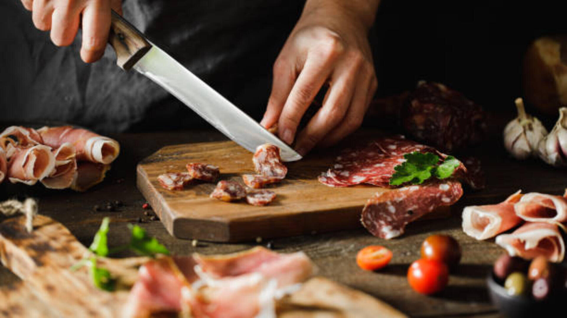 Eine Person schneidet gepökeltes Fleisch auf einem Holzbrett, umgeben von verschiedenen Fleischsorten, Knoblauch, Kirschtomaten, Oliven und Kräutern auf einem rustikalen Tisch.