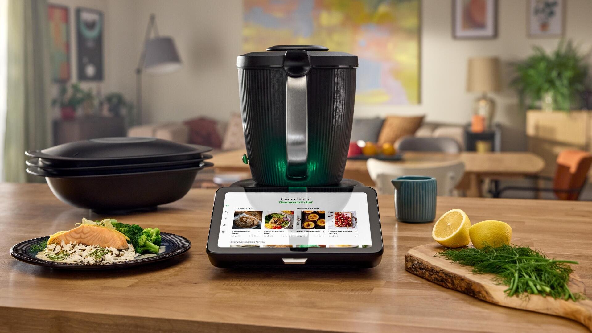 Ein intelligentes Küchengerät mit einem digitalen Bildschirm, auf dem Rezepte angezeigt werden, steht auf einer Holztheke, umgeben von Lachs, Reis, Gemüse, Zitronen, Kräutern, gestapelten Tabletts und einem blauen Becher in einer gemütlichen, modernen Küche.