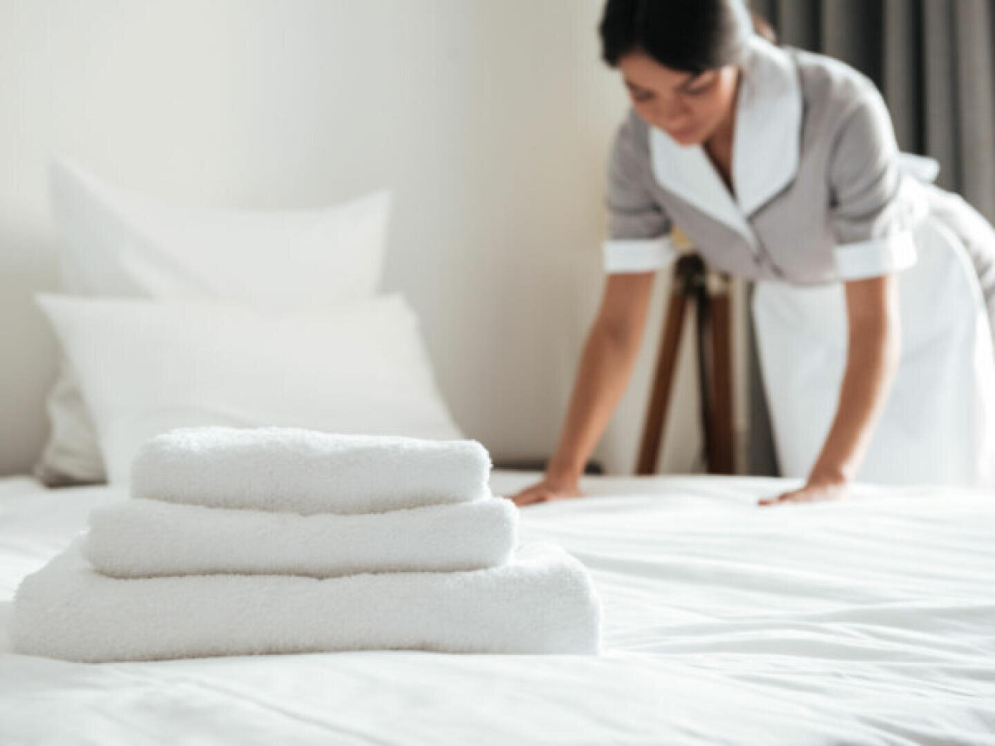 Eine Haushälterin in Hoteluniform macht ein Bett, im Vordergrund liegt ein ordentlicher Stapel weißer Handtücher. Das Zimmer sieht sauber und hell aus.