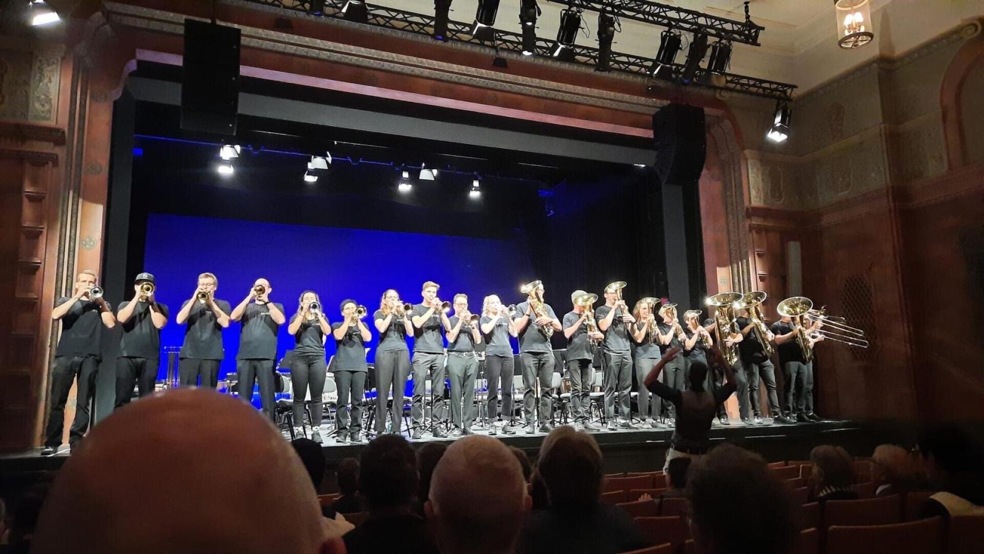 Auf der Bühne spielt eine schwarz gekleidete Blaskapelle, die in einer Reihe steht und ihre Instrumente spielt, während ein Dirigent von vorne dirigiert. Die Zuschauer sehen von ihren Plätzen in einem Theater mit verzierten Wänden zu.