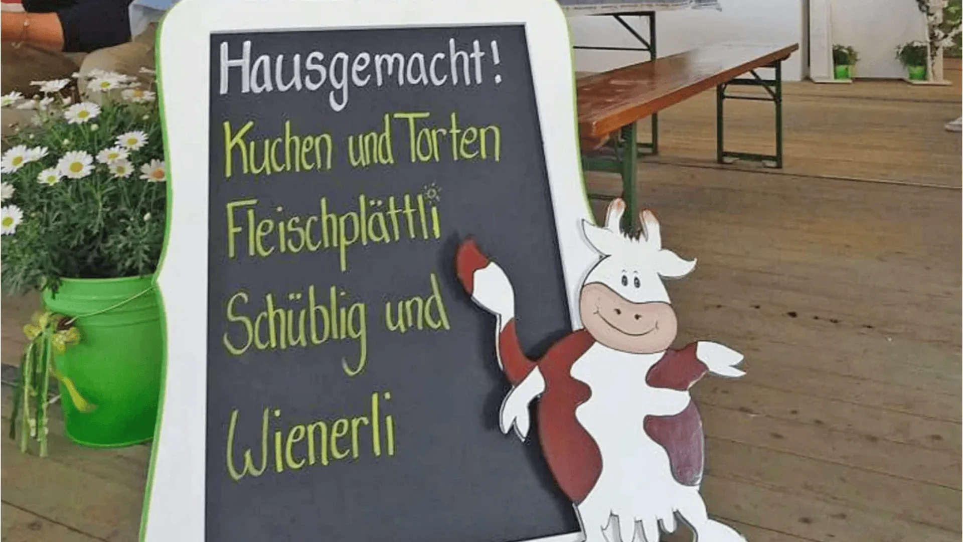 Eine Tafel in deutscher Sprache wirbt für hausgemachte Kuchen, Torten, Fleischplatten, Wurst und Wienerli. Daneben steht eine lächelnde Cartoon-Kuh, im Hintergrund ein grüner Eimer mit weißen Blumen und Holztischen.