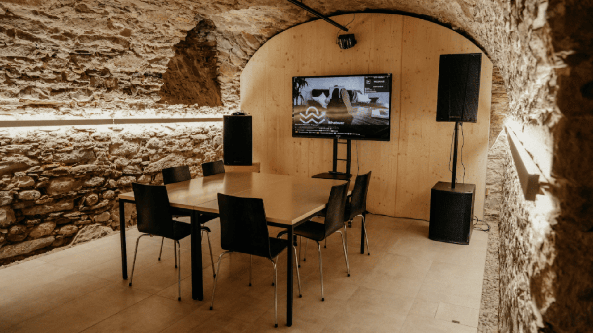 Ein moderner Besprechungsraum mit Steinwänden und gewölbter Decke, der mit einem rechteckigen Tisch mit sechs schwarzen Stühlen, einem großen Fernsehbildschirm und Lautsprechern an beiden Seiten ausgestattet ist, in einem warm beleuchteten, minimalistischen Ambiente.
