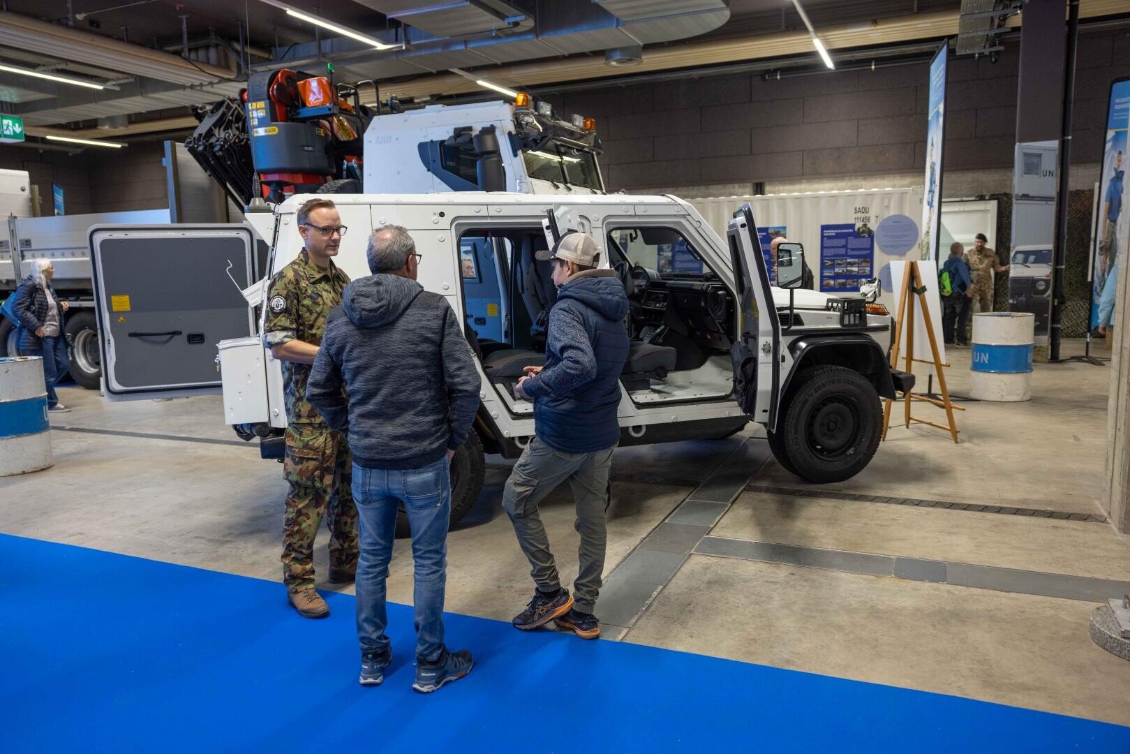 Drei Männer, darunter einer in Militäruniform, stehen und unterhalten sich neben einem weißen gepanzerten Fahrzeug mit offenen Türen in einer Ausstellungshalle. Im Hintergrund sind weitere Personen und Auslagen zu sehen.