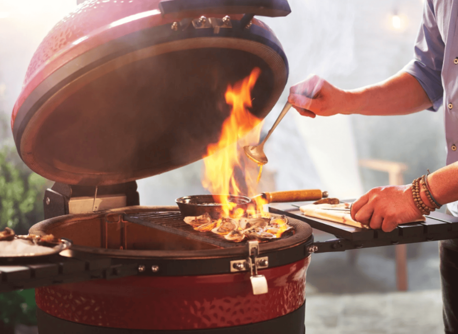Eine Person grillt Hähnchen auf einem roten Kamadogrill mit offenem Deckel und gießt mit einer Schöpfkelle Soße darüber, während die Flammen vom Grill aufsteigen. Kochutensilien und Holzbretter liegen daneben.