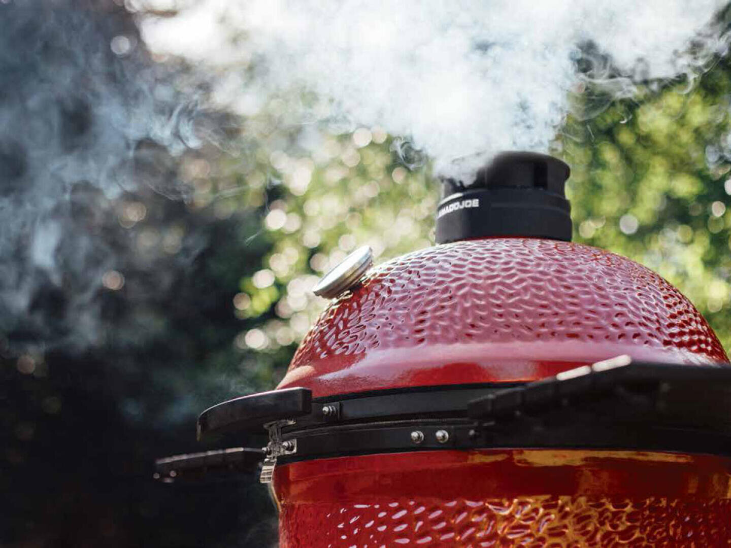 Ein roter Keramikgrill mit Rauch, der aus dem Lüftungsschacht oben aufsteigt, vor einem unscharfen, grünen Hintergrund an einem sonnigen Tag.
