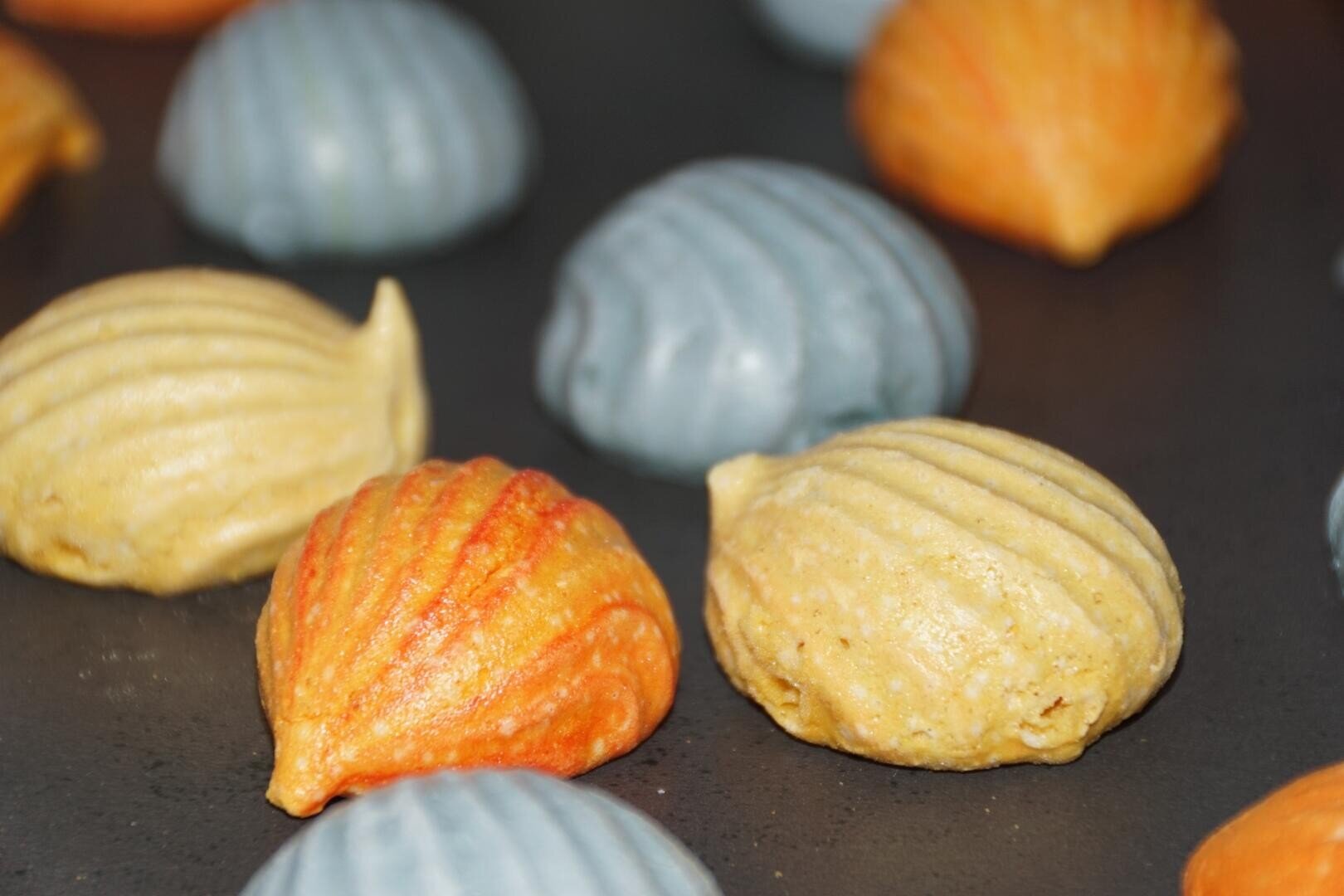 Muschelförmige Kekse in Orange-, Gelb- und Blautönen sind auf einer dunklen Oberfläche angeordnet, die an Muscheln mit Rillenmuster erinnert.
