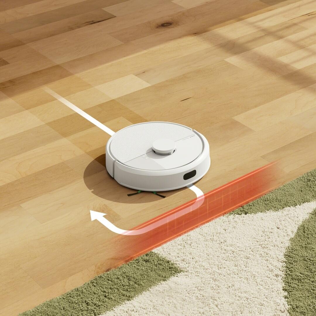 Ein weißer Staubsaugerroboter reinigt einen Holzboden und nähert sich dabei einem grün-weißen Teppich. Eine rote virtuelle Barriere und ein Pfeil zeigen an, dass der Staubsauger so programmiert ist, dass er den Teppichbereich nicht betritt.
