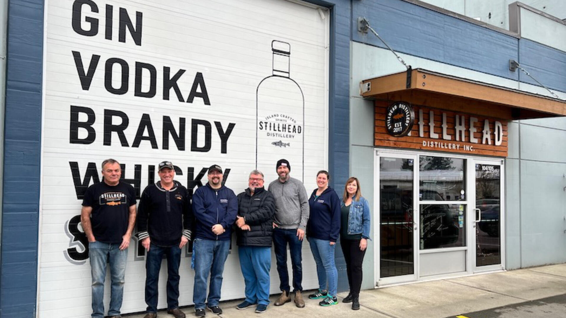 Sieben Personen stehen lächelnd vor der Stillhead Distillery, neben einem großen Schild, auf dem Gin, Wodka, Brandy und Whisky aufgelistet sind, mit der Abbildung einer Flasche und dem Eingang der Destillerie auf der rechten Seite.