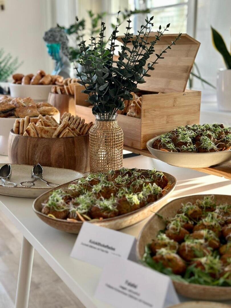 Ein gedeckter Tisch mit verschiedenen Fingerfoods und Gebäck in Holztabletts und Schalen, garniert mit Grünzeug und Microgreens. Eine Glasvase mit grünem Blattwerk schmückt die Mitte. Natürliches Licht erhellt die Szene.
