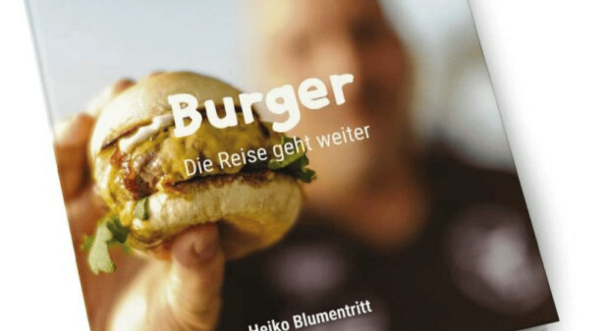 Eine Nahaufnahme einer Person, die einen Burger vor einem Buchcover mit dem Titel Burger: Die Reise geht weiter von Heiko Blumentritt. Der Burger ist scharf, während die Person und der Hintergrund unscharf sind.