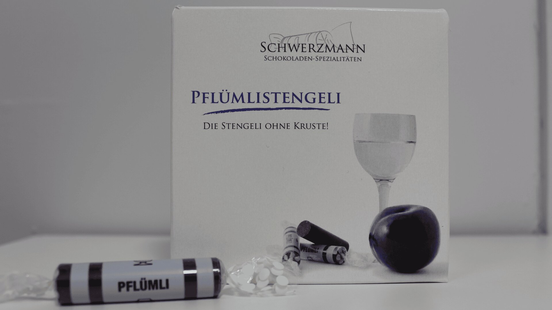 Eine Schachtel mit der Aufschrift Schwerzmann Schokoladen-Spezialitäten mit Pflümlistengeli und der Abbildung eines Weinglases, einer Pflaume und Schokoladenstäbchen. Davor befindet sich ein kleines eingewickeltes Bonbon mit der Aufschrift Pflümli.