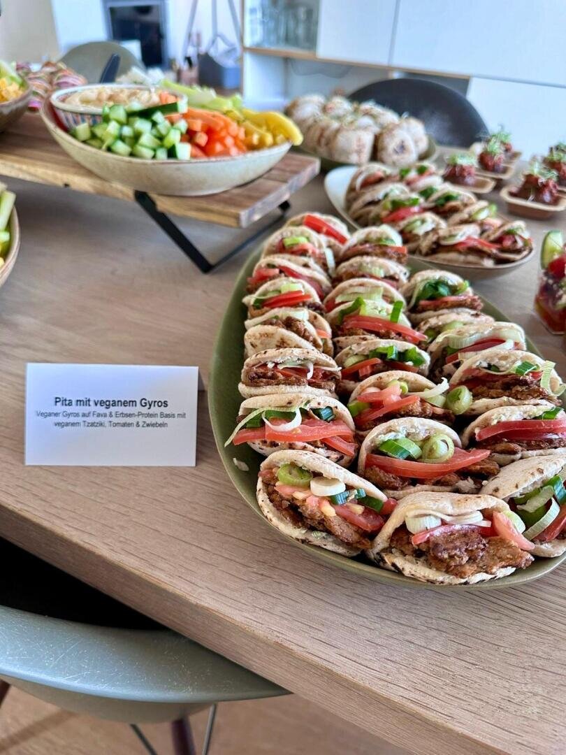 Auf einem Tisch steht ein Teller mit Fladenbrot, gefüllt mit veganem Gyros, Tomaten, Gurken und Zwiebeln. Dahinter stehen Schalen mit gehacktem Gemüse und anderen Gerichten. Ein Etikett auf Deutsch beschreibt das vegane Pita-Gyros.