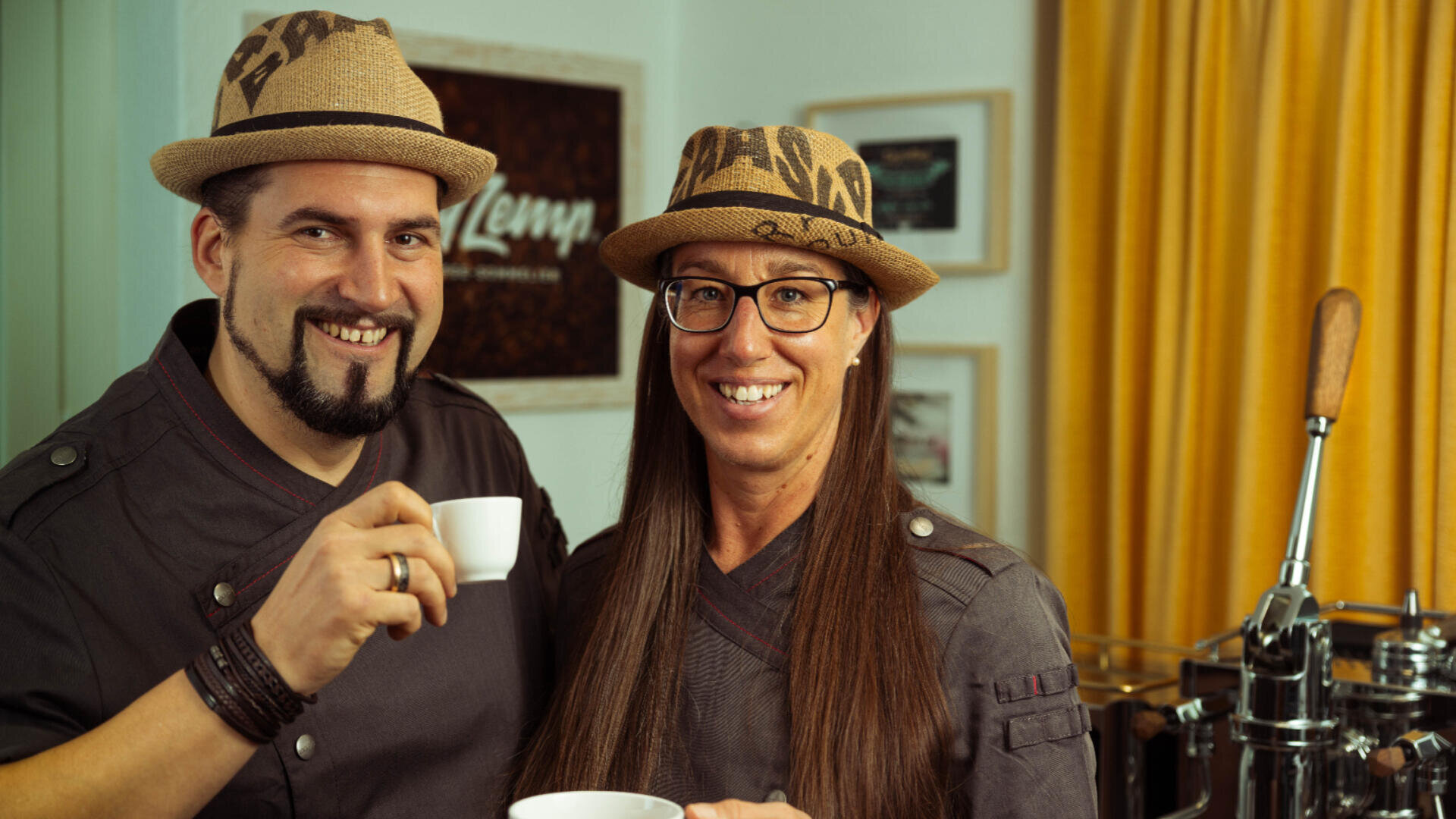 Zwei Personen in Kochjacken und Strohhüten, lächelnd und mit weißen Kaffeetassen in der Hand, stehen in einem gemütlichen Raum mit gerahmten Bildern an der Wand und gelben Vorhängen im Hintergrund.