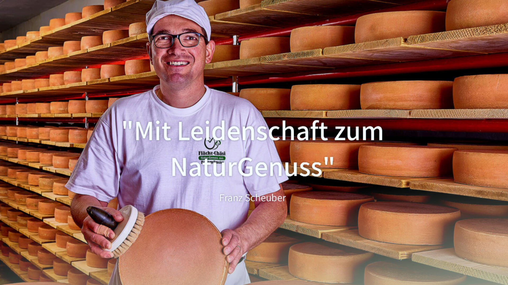 Ein lächelnder Mann in weißer Uniform und Mütze hält einen runden Käse und einen Pinsel vor Regalen, die mit Käselaiben gefüllt sind. Ein deutscher Text überlagert das Bild: "Mit Leidenschaft zum NaturGenuss" und "Franz Scheuber.".