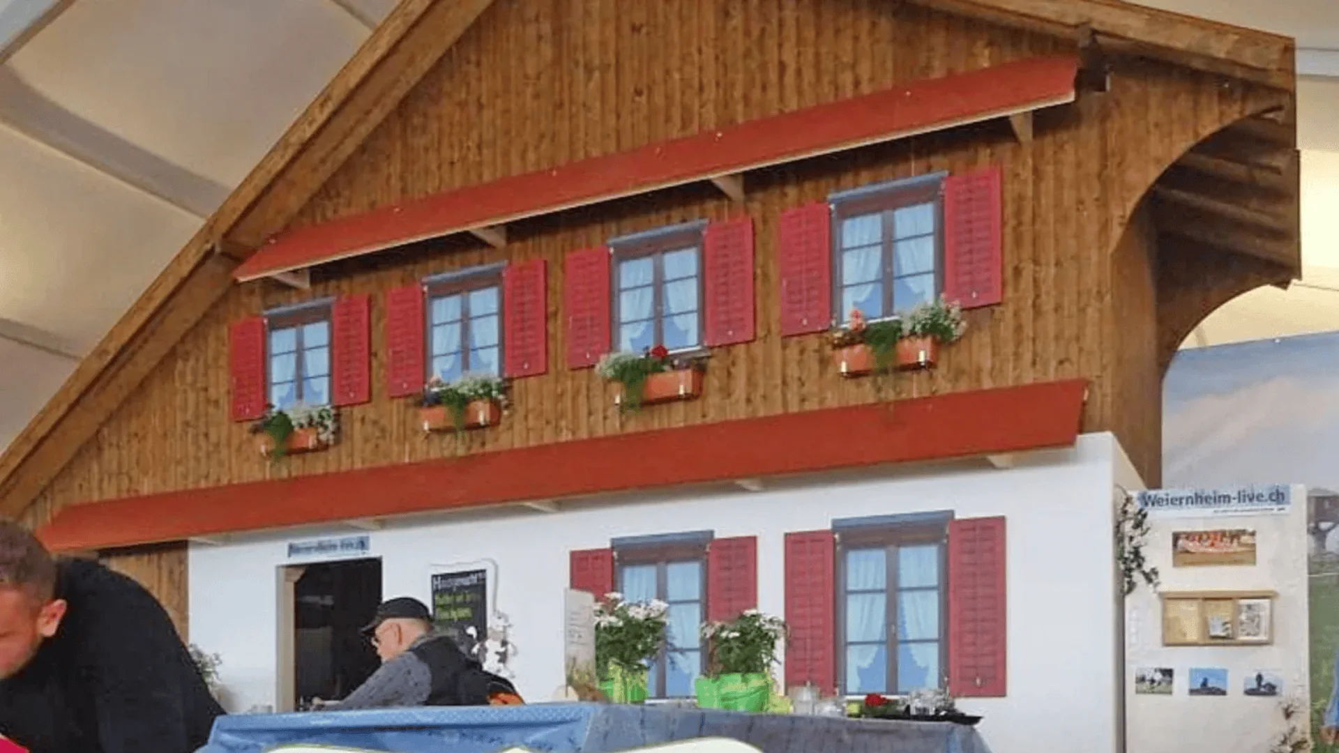 Eine Holzhausfassade mit roten Fensterläden und Blumenkästen ist im Innenbereich zu sehen. Davor sitzen Menschen an Tischen, und unter den Fenstern sind Dekorationen und Hinweisschilder zu sehen.