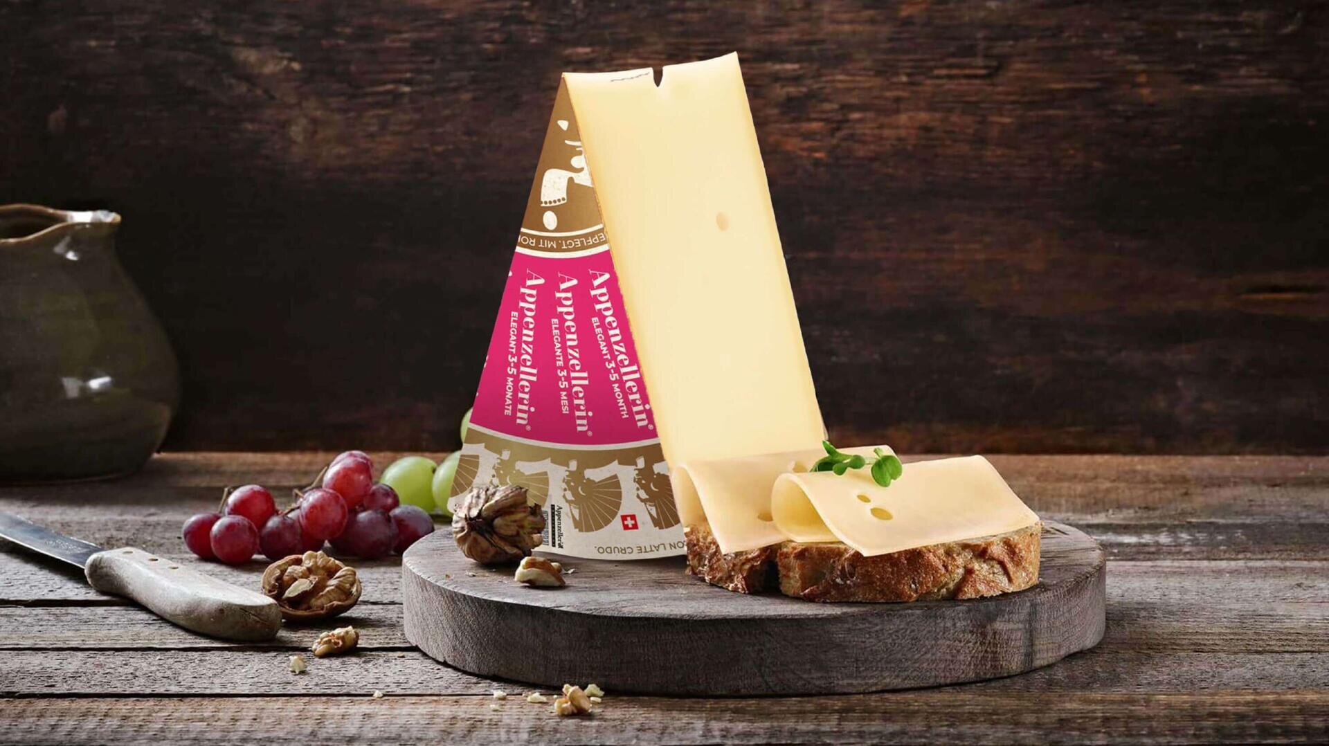 Ein Stück Appenzeller Käse steht aufrecht auf einem Holzbrett, daneben zwei Käsescheiben auf Brot, Weintrauben, Walnüsse und ein rustikaler Krug im Hintergrund.
