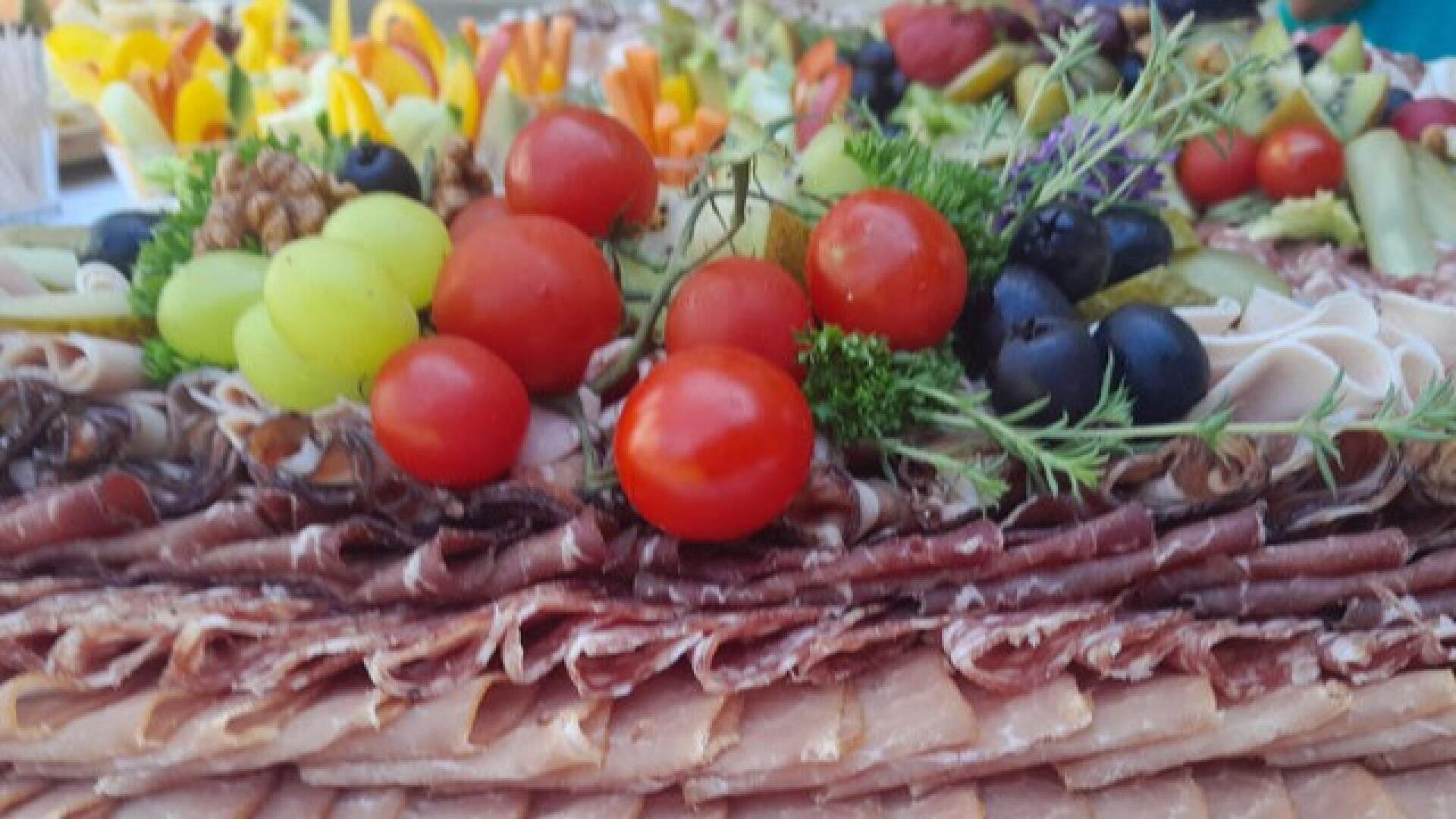 Eine Nahaufnahme einer Wurstplatte mit mehreren Schichten Wurstwaren, Kirschtomaten, Trauben, Oliven, Walnüssen und buntem Gemüse im Hintergrund.