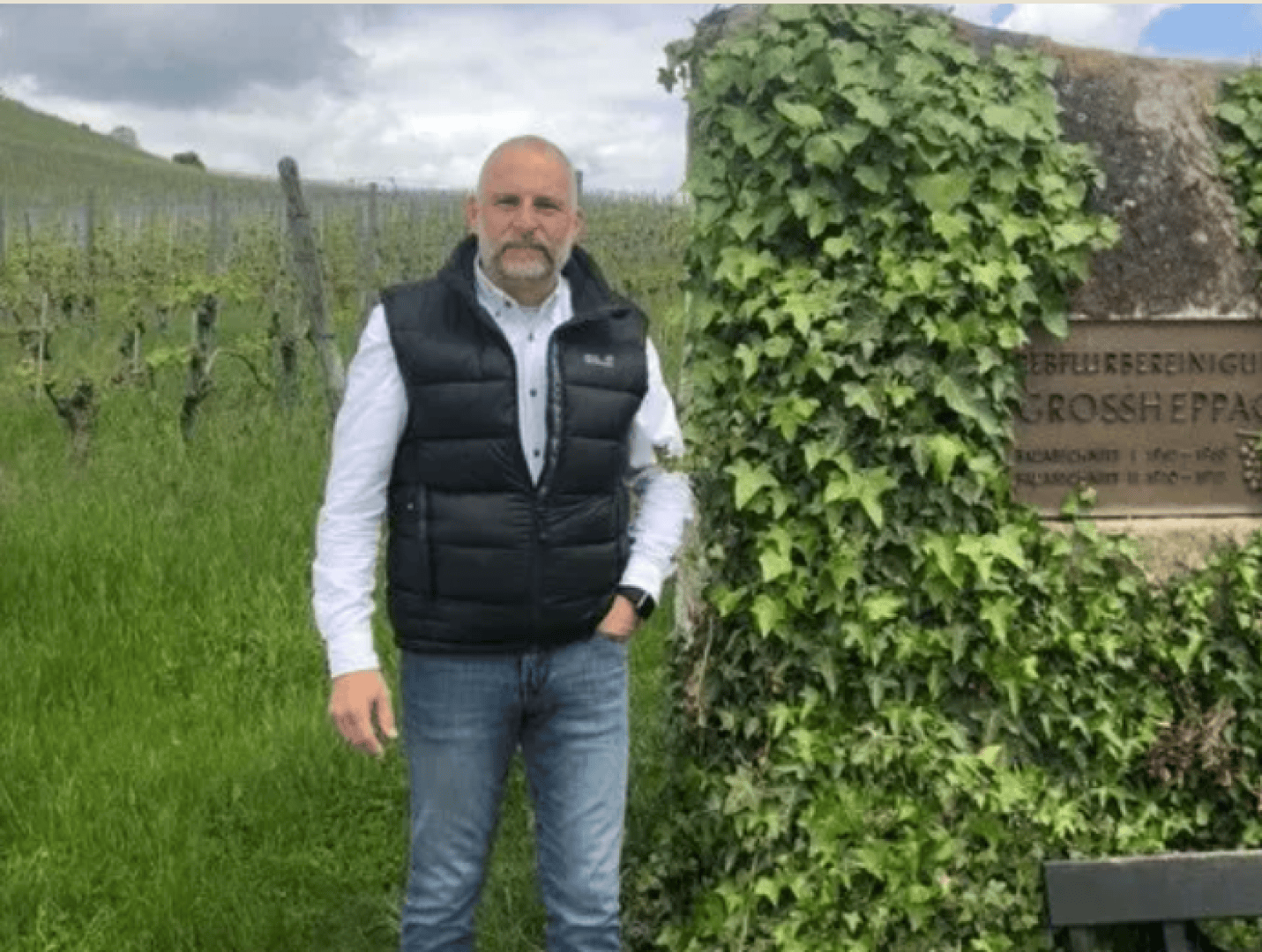 Ein Mann in schwarzer Weste und Jeans steht neben einem mit Efeu bewachsenen Stein, auf dem eine Gedenktafel teilweise sichtbar ist. Hinter ihm ist ein üppiges, grünes Feld mit Pflanzenreihen und einem wolkenverhangenen Himmel zu sehen.