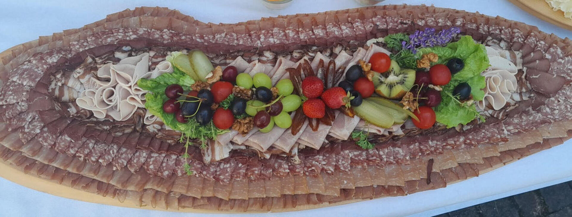 Eine Auswahl an in Scheiben geschnittenen Wurstwaren, die in konzentrischen Reihen auf einer Platte angeordnet sind, mit einer bunten Mitte aus Salat, Trauben, Erdbeeren, Tomaten, Gurken, Kiwi und essbaren Blumen.