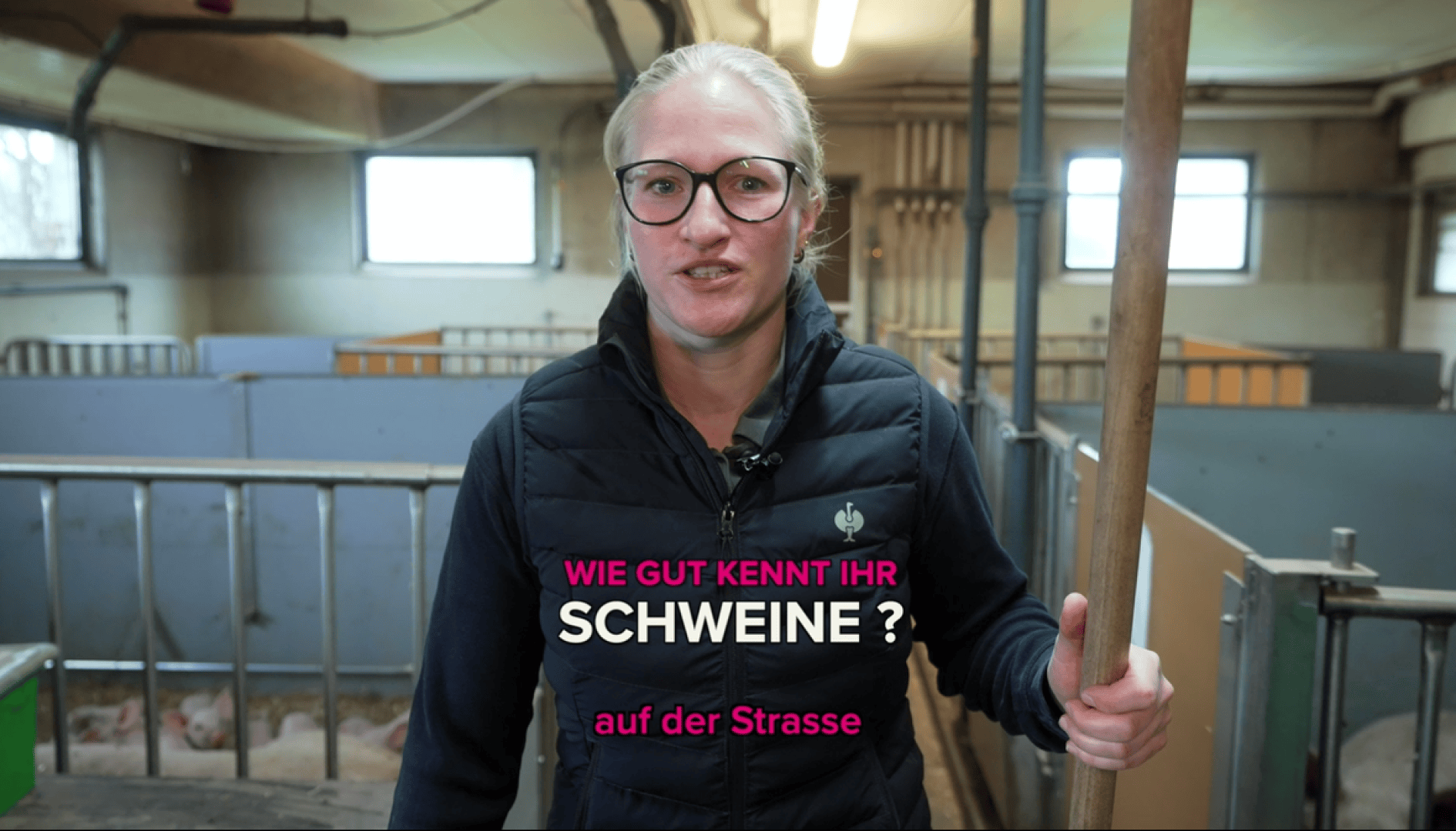 Eine Frau mit Brille und schwarzer Weste steht in einem Schweinestall und hält einen Holzstock. Der Text auf dem Bild lautet: Wie gut kennt ihr Schweine? auf der Straße. Im Hintergrund sind Schweineställe und Ferkel zu sehen.