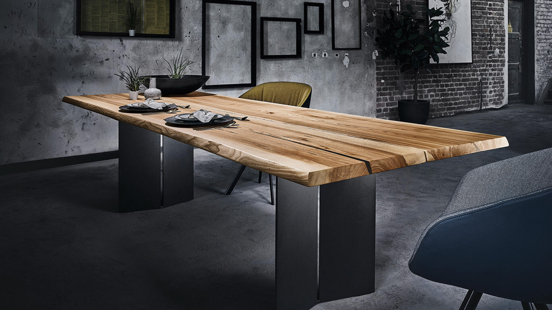 Ein modernes Esszimmer mit einem dicken Holztisch auf Metallbeinen, bestückt mit Tellern und Tassen. Der Hintergrund besteht aus grauen, strukturierten Wänden, gerahmter Kunst und freiliegenden Ziegelsteinen, mit modernen Stühlen um den Tisch.