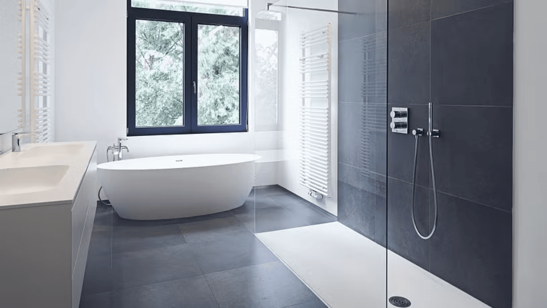 Modernes Badezimmer mit freistehender weißer Badewanne an einem großen Fenster, daneben eine geräumige Dusche mit Glasabtrennung und dunklen Fliesen sowie ein breites Waschbecken auf der linken Seite.