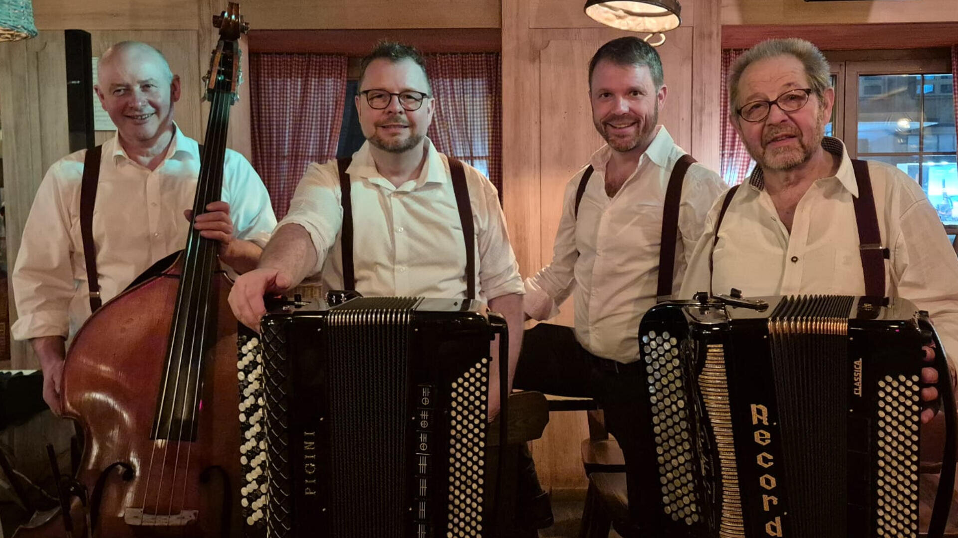 Vier Männer in weißen Hemden und Hosenträgern posieren drinnen mit Musikinstrumenten, darunter ein Kontrabass und zwei Akkordeons, und lächeln in einem warm beleuchteten, rustikalen Raum mit Holzwänden und roten Vorhängen.