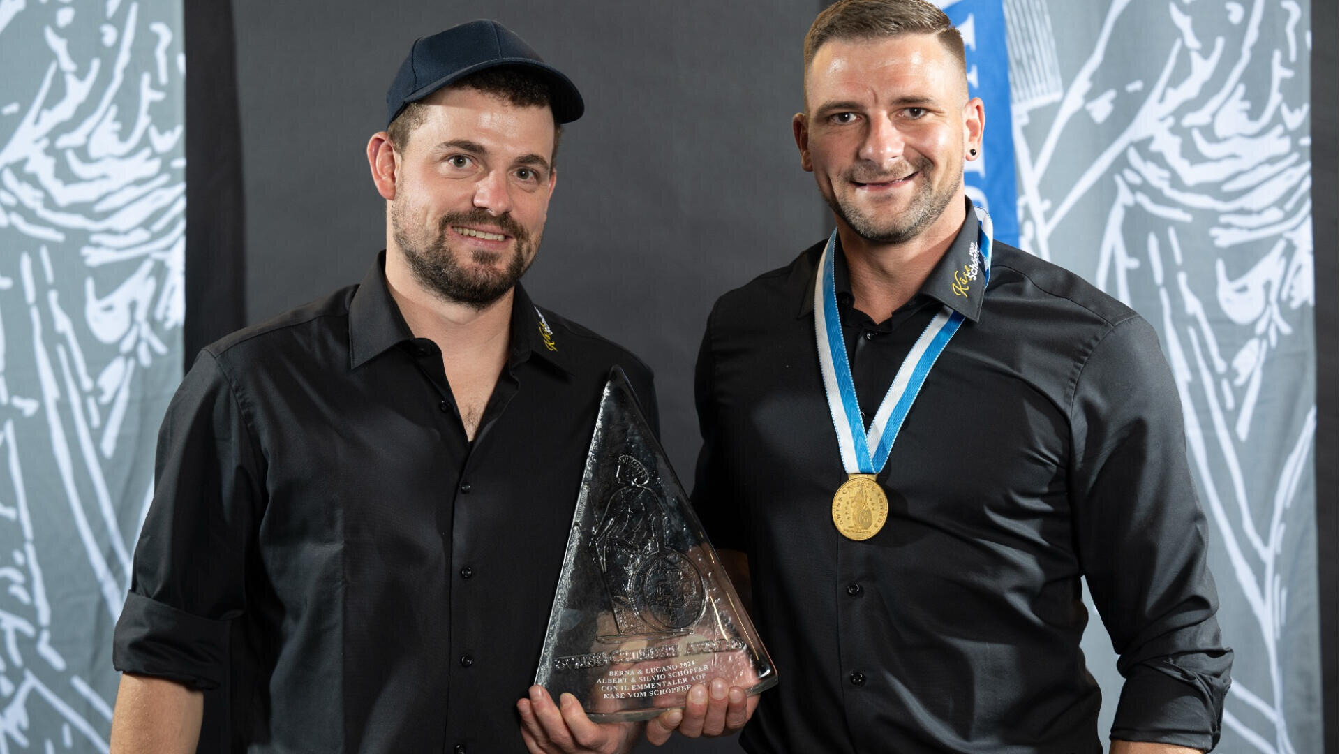 Zwei Männer in schwarzen Hemden posieren zusammen. Der eine trägt eine schwarze Mütze und hält eine große dreieckige Glasauszeichnung. Der andere hat eine blau-weiße Medaille um den Hals. Beide lächeln vor einem gemusterten Hintergrund in Graustufen.