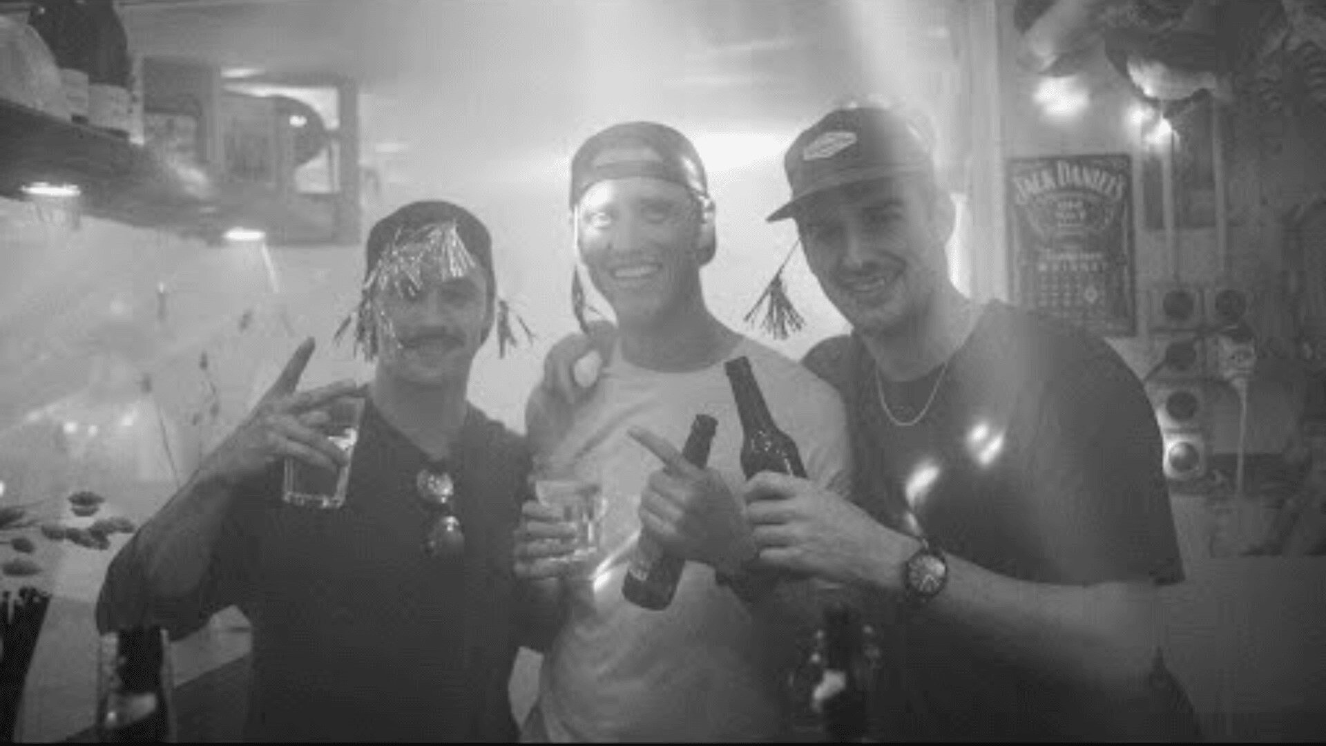Drei junge Männer lächeln auf einer Party und halten Getränke in der Hand. Zwei tragen Baseballmützen, einer hat Lametta im Haar. Die Szenerie ist schwach mit Lichtstrahlen beleuchtet, und im Hintergrund ist Partydekoration zu sehen.